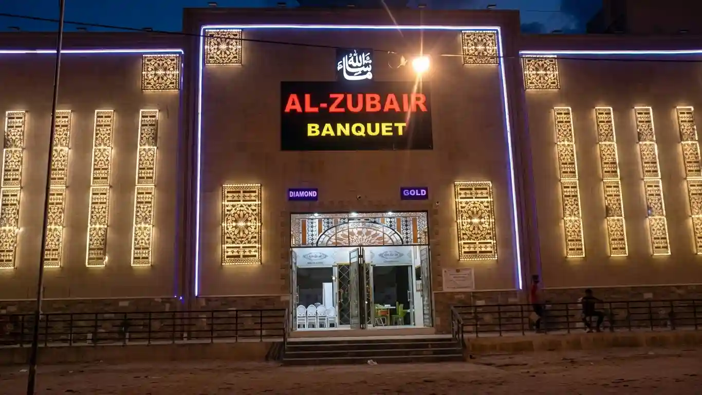 The exterior of the modern Al-Zubair Banquet hall building at night, featuring illuminated red and yellow signage, intricate geometric lattice screens (jalis) backlit with warm lights, and a set of steps leading up to the glass entrance under a deep blue hour sky.
