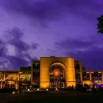 Illuminated waterfront boardwalk, warm festoon lights, and rustic Asian-style dining stalls reflecting on the Chinna Creek waters at Port Grand in Karachi at night