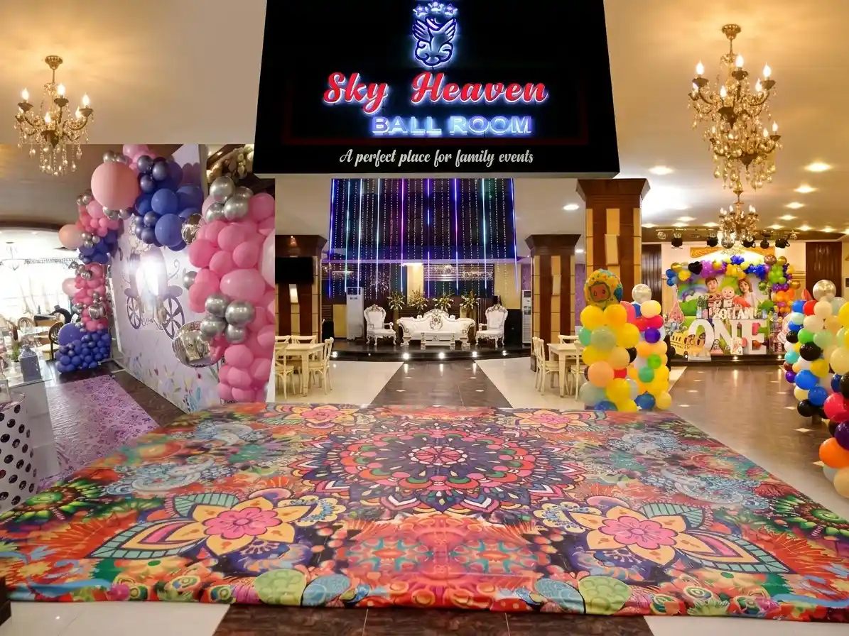 A 4-panel photo collage featuring the Sky Heaven Ballroom. Panels show: 1) The night facade with blue, red, and yellow LED string lights and an illuminated sign; 2) An interior pink and blue balloon arch with a Cinderella carriage backdrop and purple patterned floor; 3) A child's 1st birthday setup with a Cocomelon theme, balloon pillars, and a stage with a large 'ONE'; 4) A luxurious formal stage with white and gold high-backed sofas and intricate wall decor